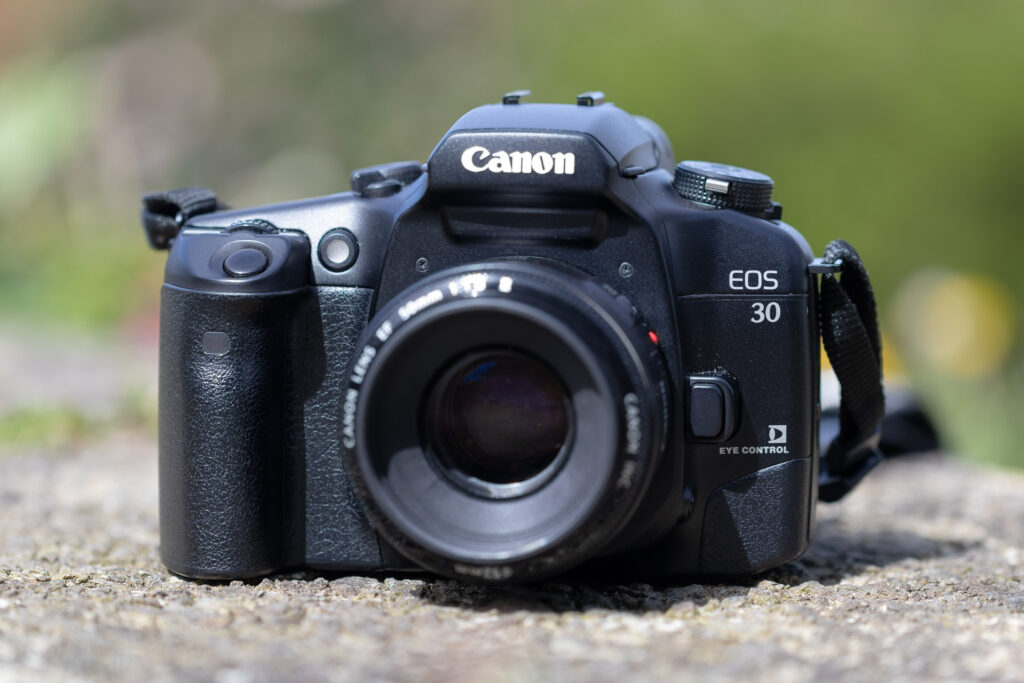 A colour photo of a Canon EOS 30 film camera, sitting on a concrete slab in an outdoor environment