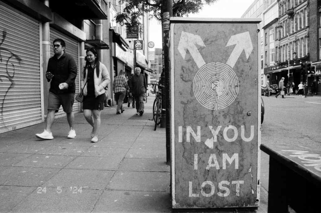 Black and white image of a couple walking down a sidewalk in Belfast. The right hand side of the frame is dominated by a sign saying "In You I Am Lost" with arrows pointing outward.