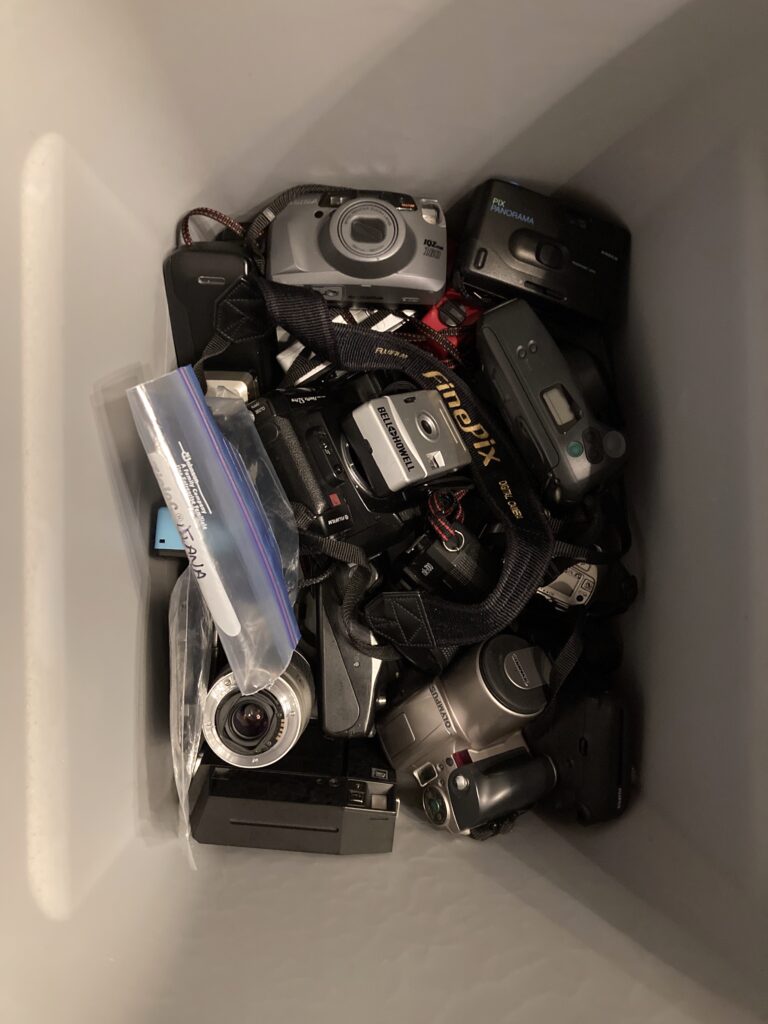 From the top of box view, a jumble of several digital and film cameras, indistinct in terms of brand, size, or function.