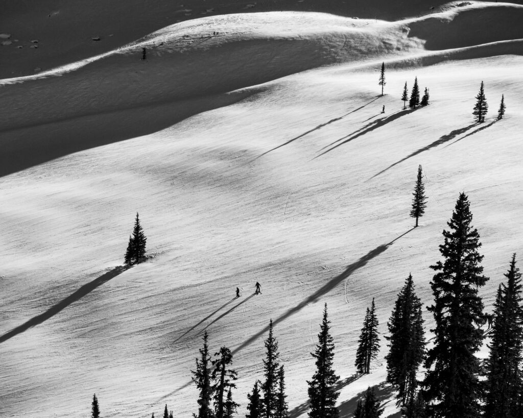 A black & white photo of a two skiers on a ski slope, with long afternoon shadows.