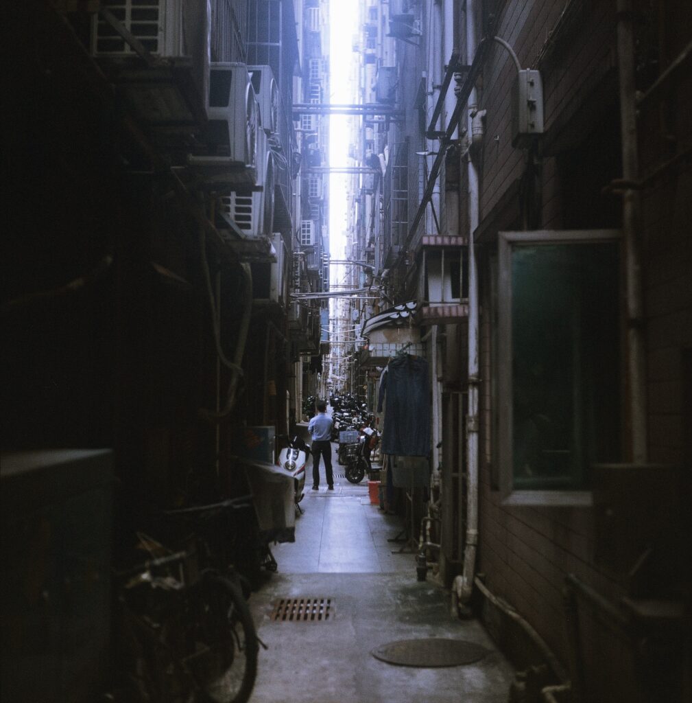 A figure stands alone in an alley between massive buildings on either side.
